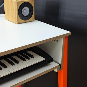 Op de afbeelding: Een wit bureau met een uitschuifbare plank met een zwart elektronisch keyboard. Het bureau heeft feloranje poten en een rand met een lichte houtnerf. Een vierkante bamboe luidspreker met een zwarte driver staat op het witte oppervlak.