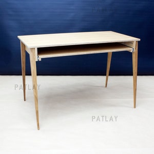 Könnte beinhalten: Ein heller Holzschreibtisch mit einer ausziehbaren Ablage. Der Schreibtisch hat vier Beine, die sich zu einer Spitze verjüngen. Der Schreibtisch steht auf einer weißen Oberfläche vor einer blauen Wand.
