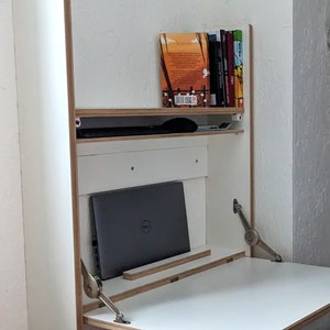 May include: White wall-mounted desk with a fold-down top and a shelf above. A laptop computer is on the desk, and a few books are on the shelf. The desk is made of plywood and has silver hinges.