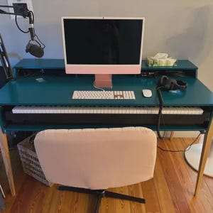 May include: A teal-colored studio desk with a built-in keyboard, a pink computer, and a white keyboard. A microphone and headphones are also visible. A light pink chair is in front of the desk.