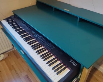 May include: A teal wooden desk with a built-in shelf and a sliding keyboard tray containing a black Roland RD-88 digital stage piano with white and black keys. The desk features a smooth, flat surface and light-coloured wooden legs.