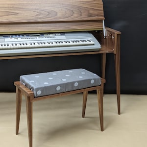 Könnte beinhalten: Ein Holzklavier mit Tastatur und passender Bank. Das Klavier hat eine dunkle Holzoberfläche und eine Klappdeckel. Die Bank hat ein graues Kissen mit weißen Punkten. Klavier und Bank stehen vor einem dunklen Hintergrund.