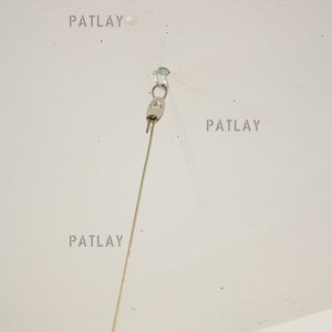 May include: A silver ceiling hook with a thin wire hanging from it. The hook is attached to a white ceiling. The word "PATLAY" is written four times on the ceiling.