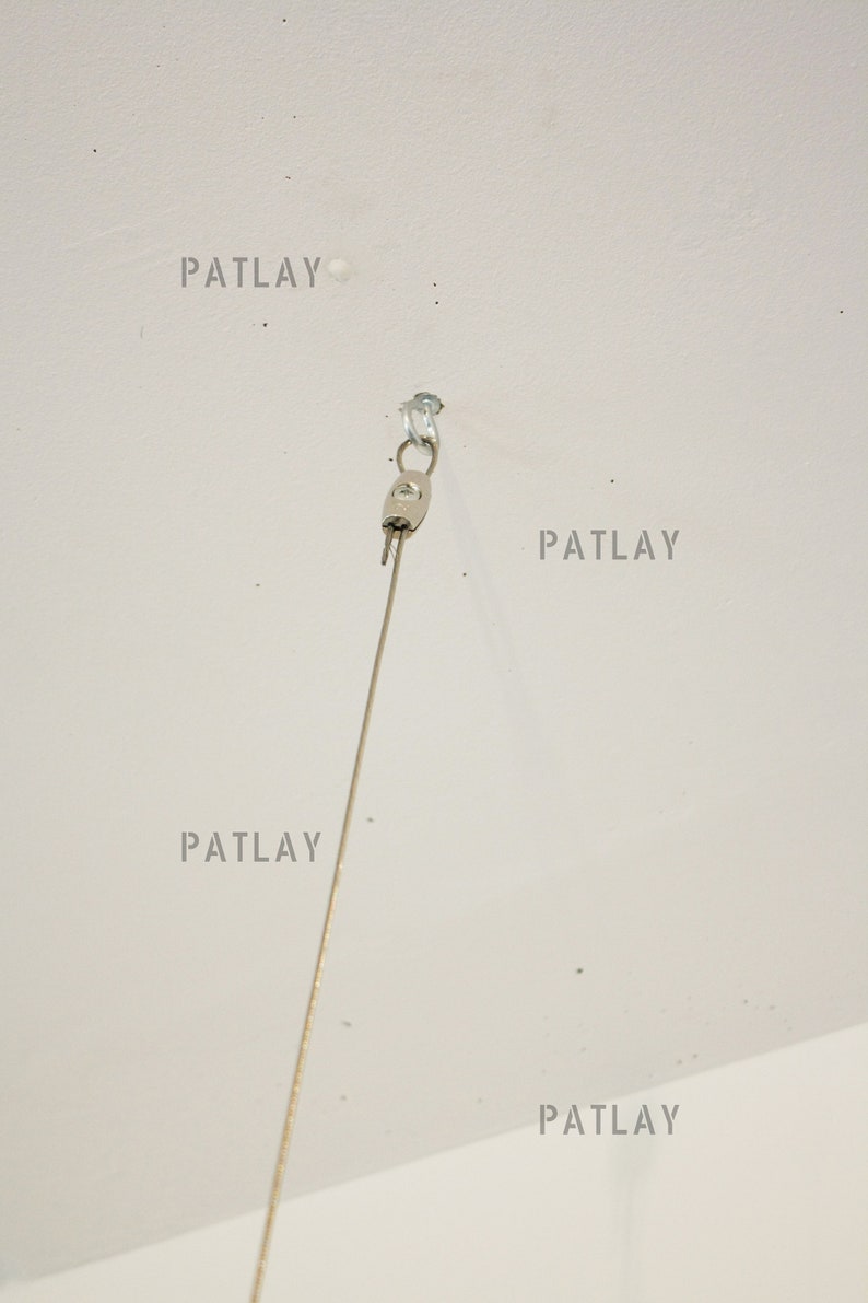 May include: A silver ceiling hook with a thin wire hanging from it. The hook is attached to a white ceiling. The word "PATLAY" is written four times on the ceiling.
