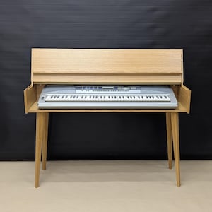 Op de afbeelding: Een lichtgekleurde houten keyboardstandaard met een ingebouwde plank. Een grijs toetsenbord staat op de plank. De standaard heeft vier schuine poten en een achterpaneel. De achtergrond is zwart.