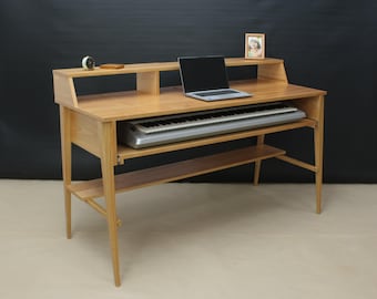 May include: A light-toned wooden studio desk featuring a raised upper shelf, a central workspace surface, and a lower horizontal support shelf. A silver electronic keyboard is housed in a sliding tray beneath the main desk surface.
