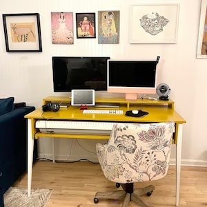 May include: A yellow desk with white legs and a white keyboard. A floral patterned office chair is in front of the desk. Two computer monitors and a tablet are on the desk. There are framed pictures on the wall behind the desk.