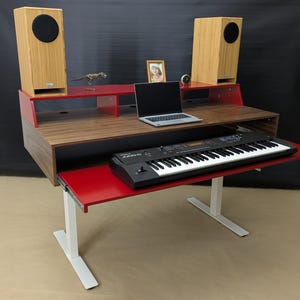 May include: A modern studio desk with a pull-out keyboard tray. The desk features a walnut finish with red accents and white legs. Two wooden speakers sit atop the desk, flanking a laptop. A black keyboard is on the pull-out tray.