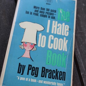Peut inclure: Un livre de poche vintage intitulé The I Hate to Cook Book de Peg Bracken avec une couverture bleue illustrée d'un personnage portant une toque de chef blanche. Le texte indique More than 180 quick and easy recipes et une citation de Jean Kerr.