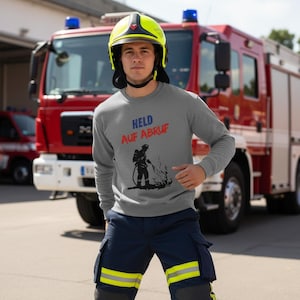 May include: A grey sweatshirt featuring the words "HELD AUF ABRUF" in blue and red, and a black graphic of a firefighter. The person is wearing a yellow helmet and navy blue trousers with yellow stripes. A red fire engine is in the background.