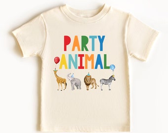 May include: A cream coloured t-shirt with the words "PARTY ANIMAL" in rainbow colours. The shirt features a giraffe, elephant, lion, and zebra wearing party hats.