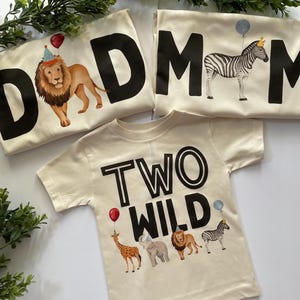 May include: Two beige t-shirts with black text. One shirt says "DAD" with a lion wearing a party hat and the other says "MOM" with a zebra wearing a crown. The third shirt is beige with black text that says "TWO WILD" and features a giraffe, elephant, lion, and zebra.