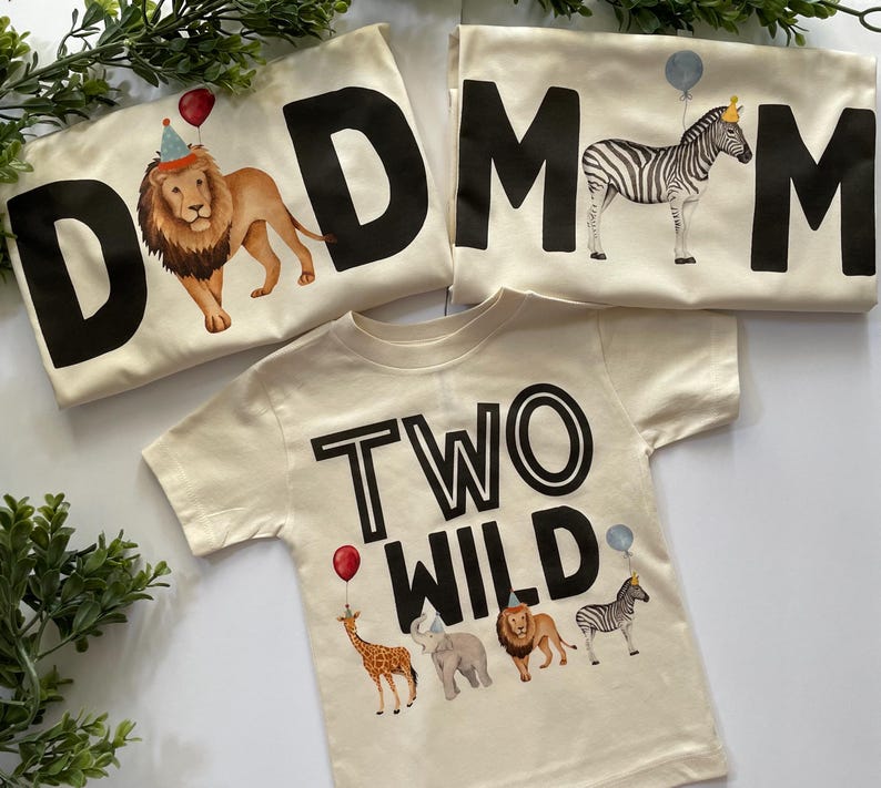 May include: Two beige t-shirts with black text. One shirt says "DAD" with a lion wearing a party hat and the other says "MOM" with a zebra wearing a crown. The third shirt is beige with black text that says "TWO WILD" and features a giraffe, elephant, lion, and zebra.