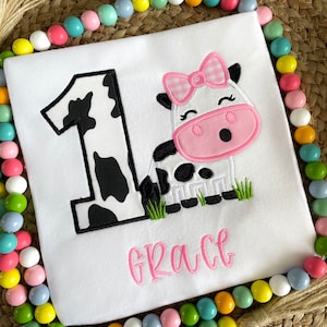 Op de afbeelding: Een witte romper met een zwart-witte koe-print nummer &eacute;&eacute;n en een roze koe met een roze strik en de naam "Grace" geborduurd in roze.