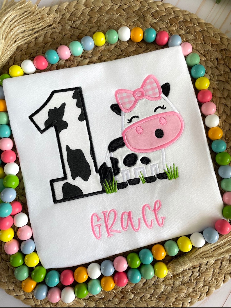 Op de afbeelding: Een witte romper met een zwart-witte koe-print nummer &eacute;&eacute;n en een roze koe met een roze strik en de naam "Grace" geborduurd in roze.