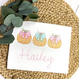 Peut inclure: Chemise blanche brod&eacute;e avec trois citrouilles orange avec des n&oelig;uds roses et bleus. Le nom "Hailey" est brod&eacute; sous les citrouilles.