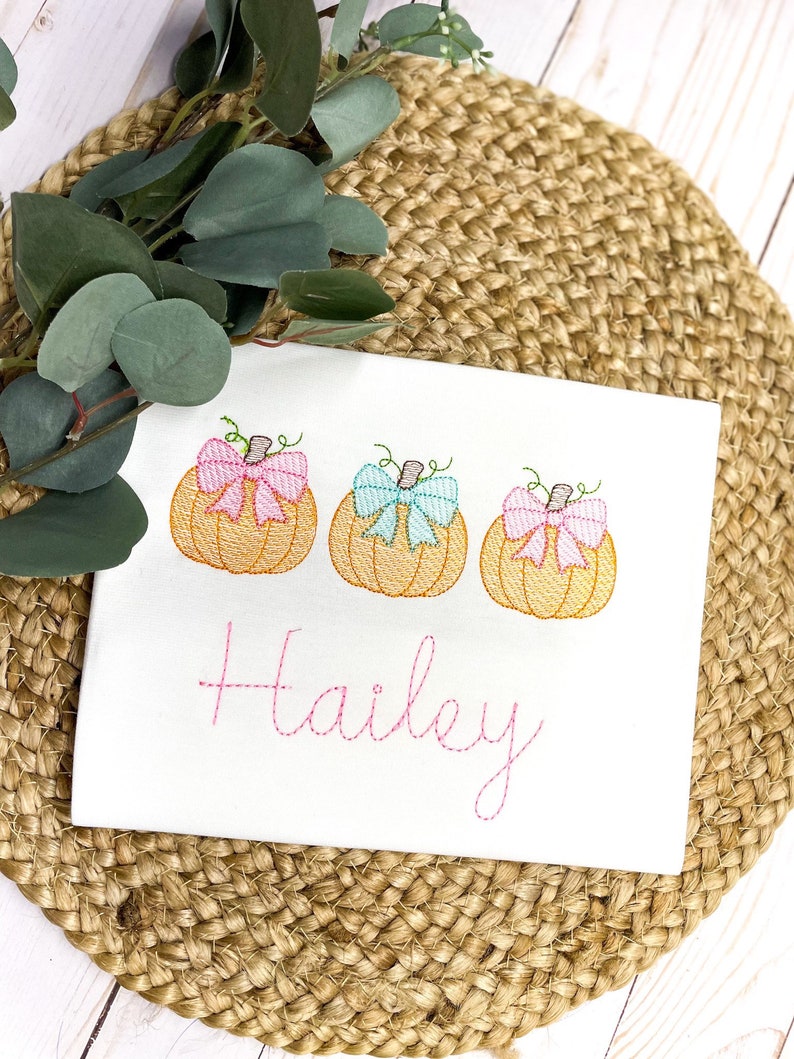 Peut inclure: Chemise blanche brod&eacute;e avec trois citrouilles orange avec des n&oelig;uds roses et bleus. Le nom "Hailey" est brod&eacute; sous les citrouilles.