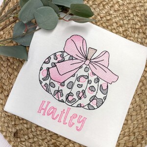 Peut inclure: Un t-shirt blanc avec une citrouille &agrave; imprim&eacute; l&eacute;opard rose et gris avec un n&oelig;ud rose. Le nom "Hailey" est brod&eacute; en rose sous la citrouille.