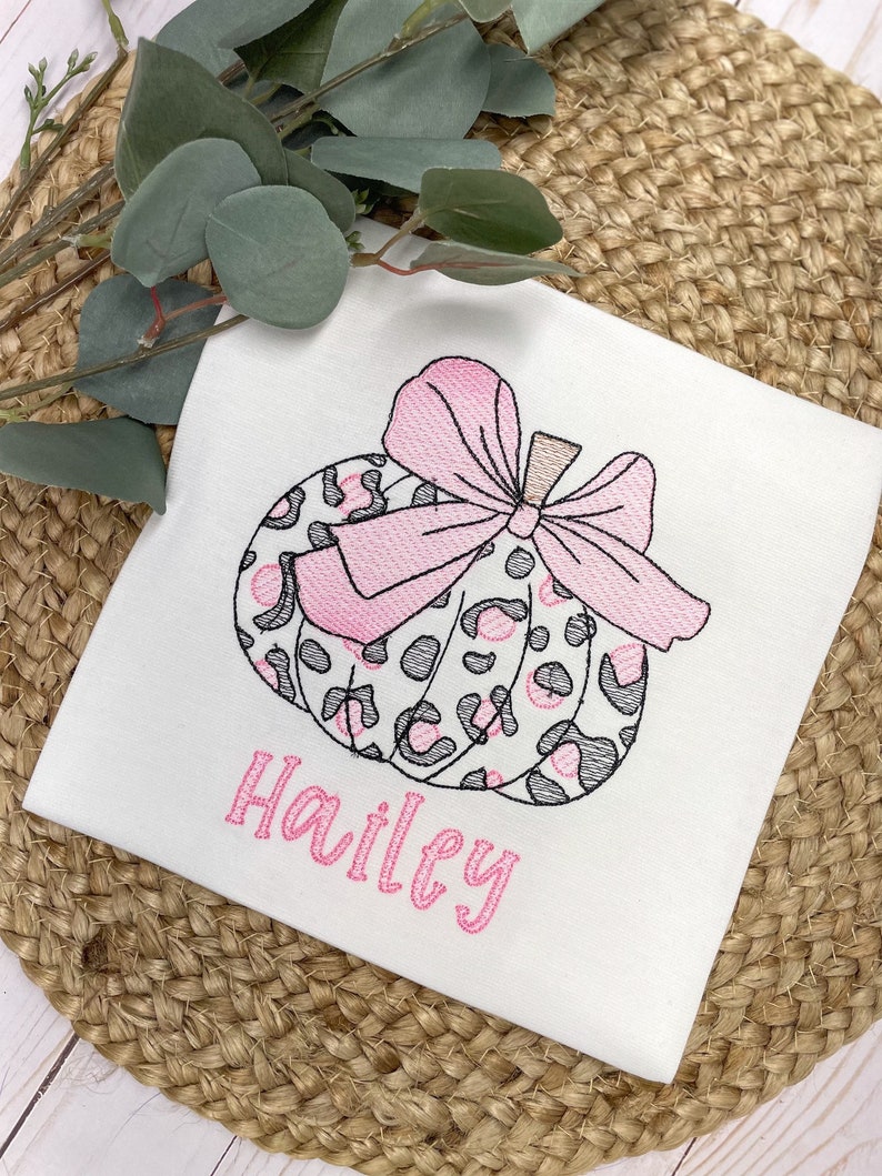 Peut inclure: Un t-shirt blanc avec une citrouille &agrave; imprim&eacute; l&eacute;opard rose et gris avec un n&oelig;ud rose. Le nom "Hailey" est brod&eacute; en rose sous la citrouille.
