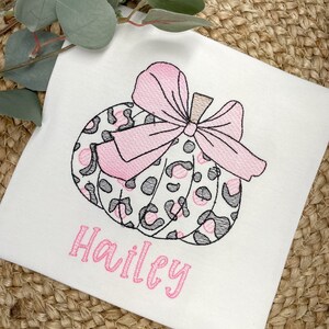 Peut inclure: Un t-shirt blanc avec une citrouille brod&eacute;e rose et noire avec un n&oelig;ud. Le nom "Hailey" est brod&eacute; sous la citrouille.