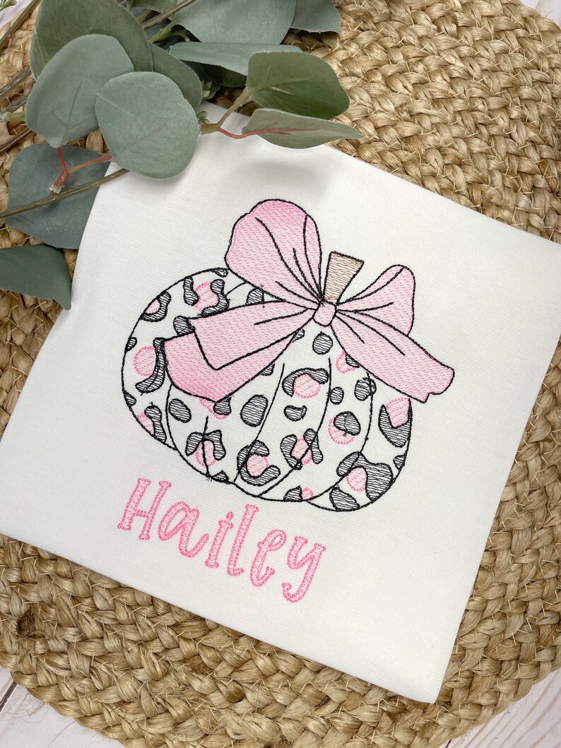 Peut inclure: Un t-shirt blanc avec une citrouille brod&eacute;e rose et noire avec un n&oelig;ud. Le nom "Hailey" est brod&eacute; sous la citrouille.