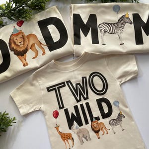 May include: Two beige t-shirts with black text and animal illustrations. One shirt says "DAD" with a lion wearing a party hat. The other shirt says "MOM" with a zebra wearing a crown. The third shirt says "TWO WILD" with a giraffe, elephant, lion, and zebra.