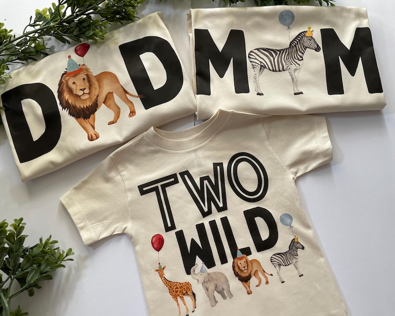 May include: Two beige t-shirts with black text and animal illustrations. One shirt says "DAD" with a lion wearing a party hat. The other shirt says "MOM" with a zebra wearing a crown. The third shirt says "TWO WILD" with a giraffe, elephant, lion, and zebra.