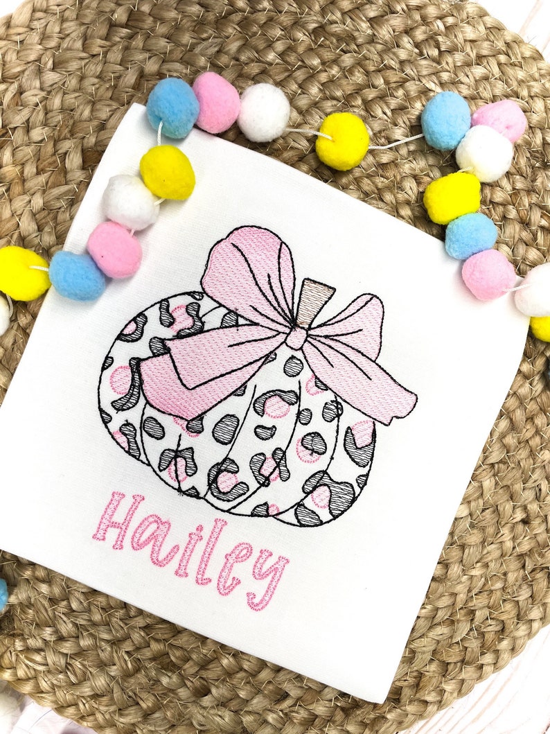 Peut inclure: Un t-shirt blanc avec une citrouille brod&eacute;e rose et grise avec un n&oelig;ud rose. Le nom "Hailey" est brod&eacute; en rose sous la citrouille.