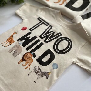 May include: A cream colored t-shirt with the text "TWO WILD" in black. The shirt features a giraffe, elephant, lion, and zebra with party hats.