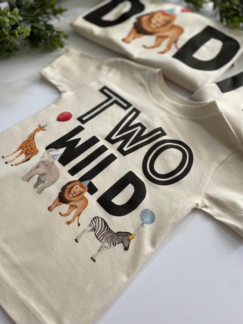 May include: A cream colored t-shirt with the text "TWO WILD" in black. The shirt features a giraffe, elephant, lion, and zebra with party hats.
