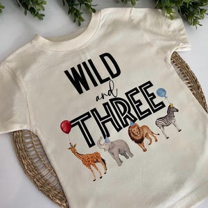 May include: A white t-shirt with the text "WILD and THREE" in black with a black outline. The shirt features a giraffe, elephant, lion, and zebra with party hats.