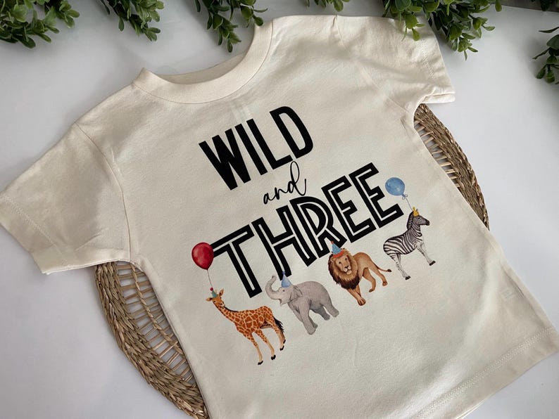 May include: A white t-shirt with the text "WILD and THREE" in black with a black outline. The shirt features a giraffe, elephant, lion, and zebra with party hats.