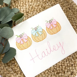 Peut inclure: Combinaison blanche avec trois citrouilles brod&eacute;es avec des n&oelig;uds roses et bleus. Le nom "Hailey" est brod&eacute; en rose sous les citrouilles.