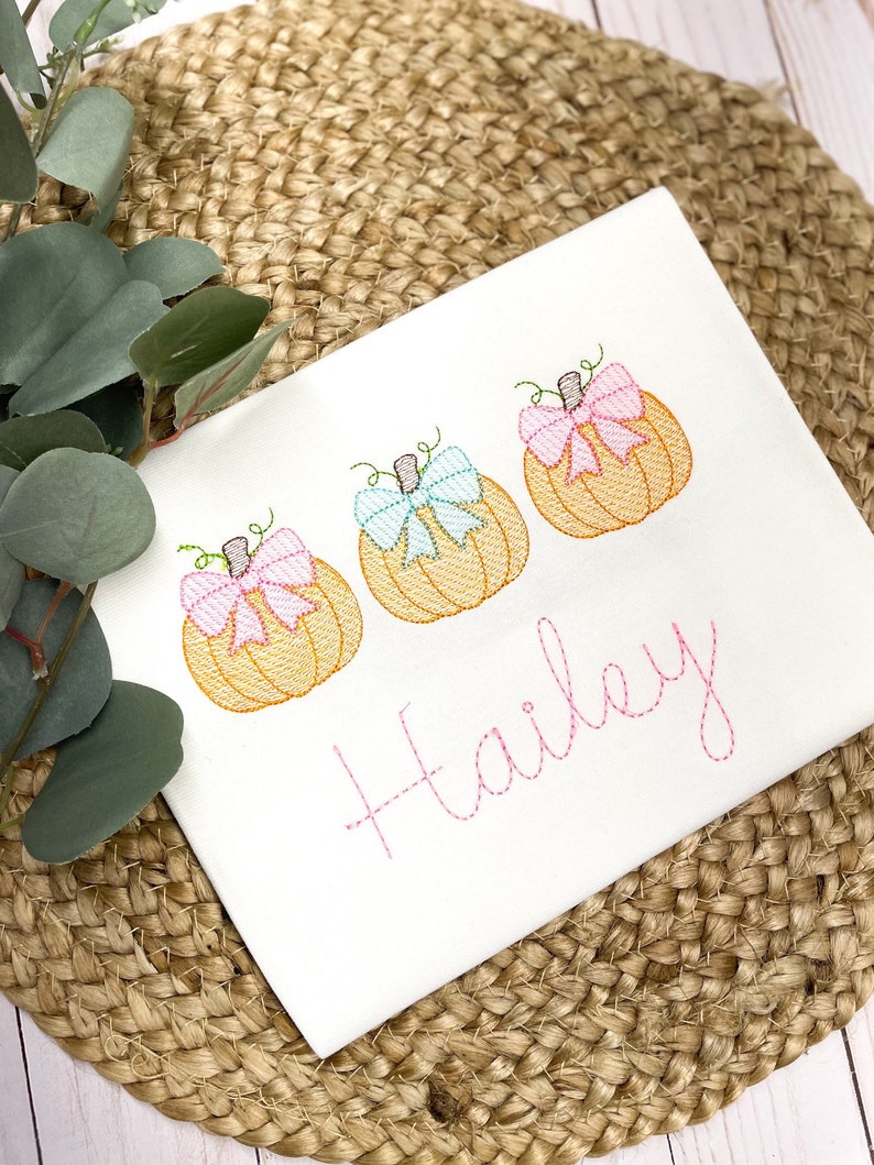 Peut inclure: Combinaison blanche avec trois citrouilles brod&eacute;es avec des n&oelig;uds roses et bleus. Le nom "Hailey" est brod&eacute; en rose sous les citrouilles.