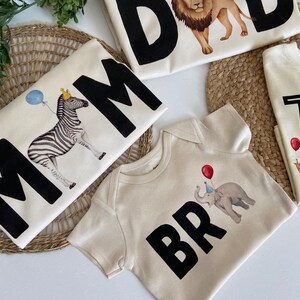 May include: Set of four cream colored t-shirts with black lettering and animal illustrations. The shirts say "Mom", "Dad", "Bro", and "Two Wild".