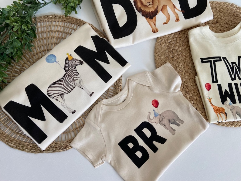 May include: Set of four cream colored t-shirts with black lettering and animal illustrations. The shirts say "Mom", "Dad", "Bro", and "Two Wild".