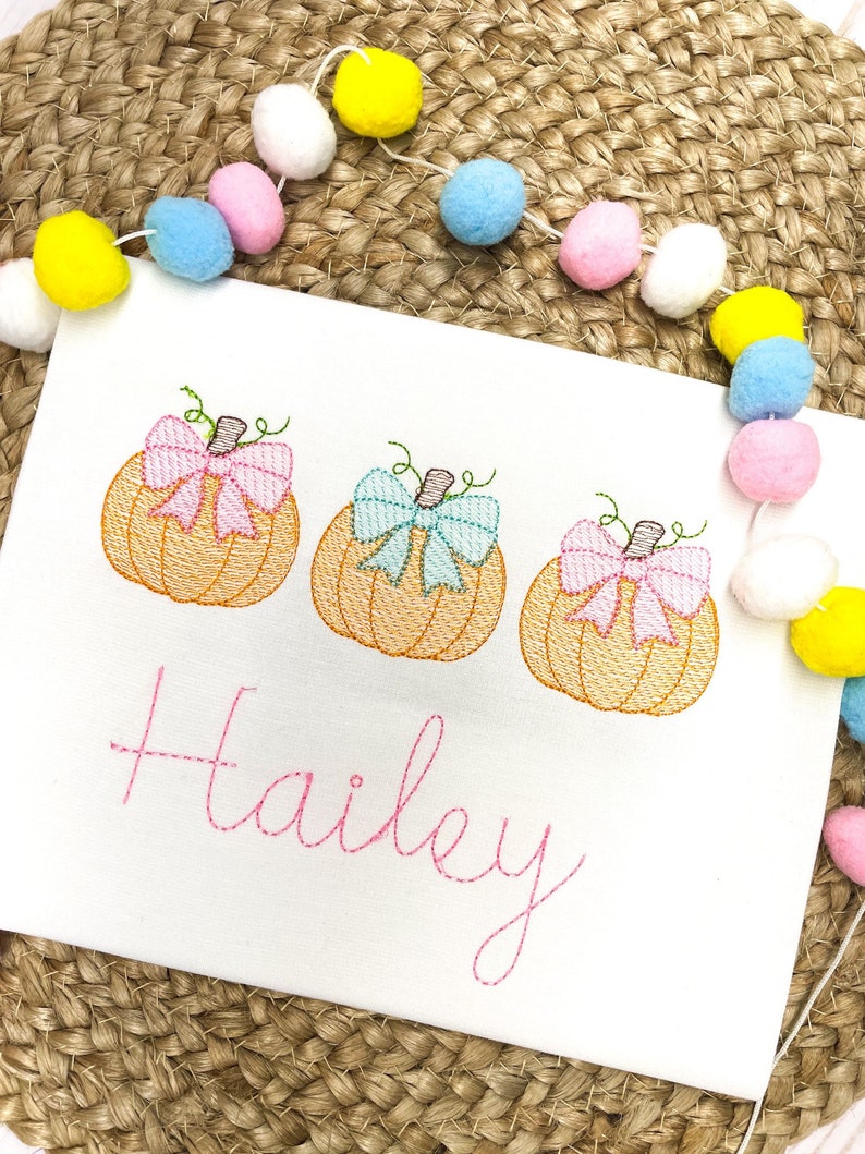 Peut inclure: Une grenouill&egrave;re blanche avec trois citrouilles brod&eacute;es avec des n&oelig;uds roses, bleus et rose p&acirc;le. Le nom "Hailey" est brod&eacute; en rose sous les citrouilles.