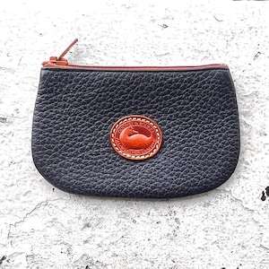 May include: A small, navy blue leather coin purse with a brown zipper and trim. The purse features a circular, brown leather patch with a duck logo. The purse is on a white, textured surface.