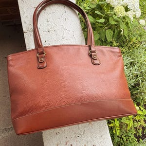 May include: A medium-brown leather tote bag with a pebbled texture on the main body and a smooth leather base panel. The bag features two matching leather shoulder straps attached with brass-toned metal hardware and a top zipper closure.
