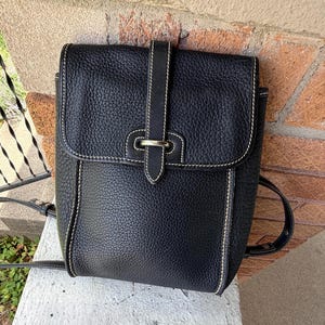 May include: A black pebbled leather backpack featuring a fold-over flap with a vertical strap and a silver-toned metal buckle closure. The bag is accented with white contrast stitching along the edges and includes thin adjustable shoulder straps.