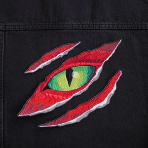 Peut inclure: Un patch brodé d'un œil déchiré en rouge et vert, cousu sur une veste en jean noire.