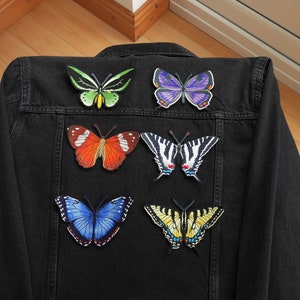 Könnte beinhalten: Eine schwarze Jeansjacke mit sieben gestickten Schmetterlings-Patches. Die Schmetterlinge sind in verschiedenen Farben, darunter Grün, Orange, Lila, Schwarz, Weiß und Gelb.