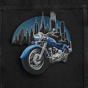 May include: An embroidered patch featuring a motorcycle in front of a city skyline. The motorcycle is blue and silver and has a large front wheel and a detailed engine.