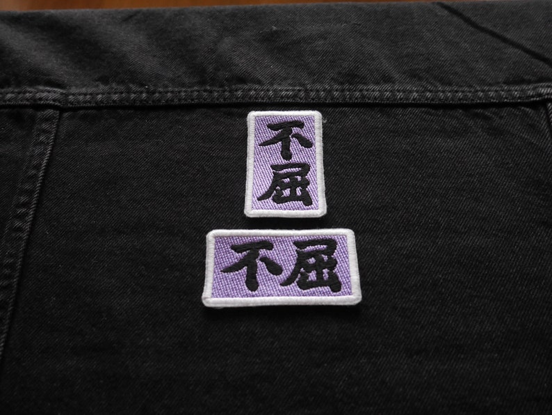 K&ouml;nnte beinhalten: Zwei violette gestickte Patches mit den chinesischen Schriftzeichen "不屈" (bedeutet "unbeugsam") in Schwarz.