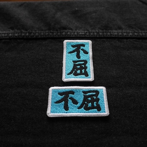 K&ouml;nnte beinhalten: Zwei blaue gestickte Patches mit wei&szlig;en Umrandungen auf einem schwarzen Jeansstoff. Die Patches zeigen die chinesischen Schriftzeichen f&uuml;r "nicht" und "schlecht" in Schwarz.