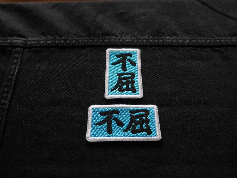 K&ouml;nnte beinhalten: Zwei blaue gestickte Patches mit wei&szlig;en Umrandungen auf einem schwarzen Jeansstoff. Die Patches zeigen die chinesischen Schriftzeichen f&uuml;r "nicht" und "schlecht" in Schwarz.