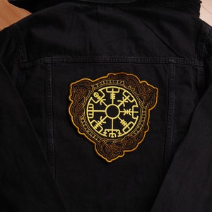 Könnte beinhalten: Eine schwarze Jeansjacke mit einem gelben gestickten Aufnäher einer Kompass mit einem Knotenmuster.