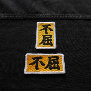 K&ouml;nnte beinhalten: Zwei gelbe gestickte Aufn&auml;her mit schwarzem Text, der "不屈" auf einer schwarzen Jeansjacke lautet.