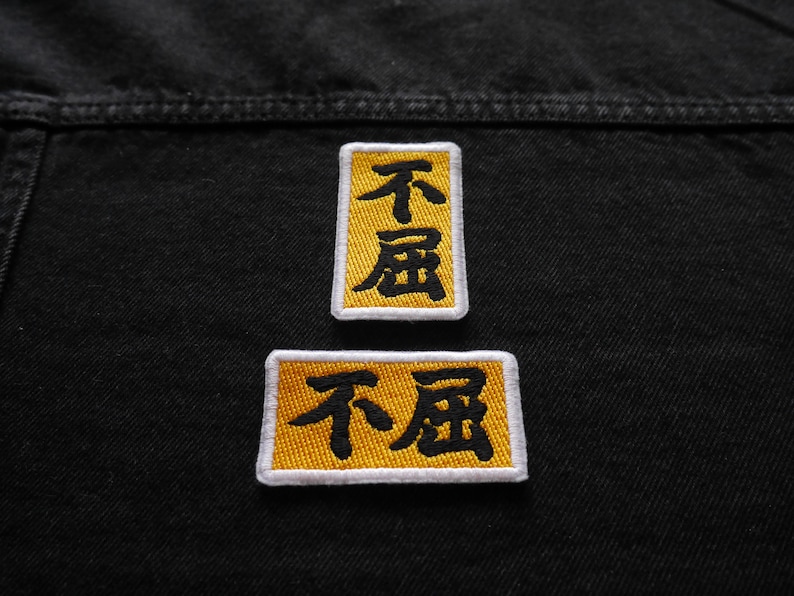 K&ouml;nnte beinhalten: Zwei gelbe gestickte Aufn&auml;her mit schwarzem Text, der "不屈" auf einer schwarzen Jeansjacke lautet.