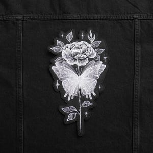 May include: White embroidered patch of a butterfly and flower on a black denim jacket.